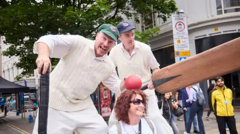 Two men on stilts in cricket gear pose with a large cricket ball on a woman's head