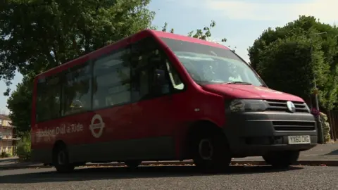 BBC A red Dial a ride minibus driving on the road with trees in the background