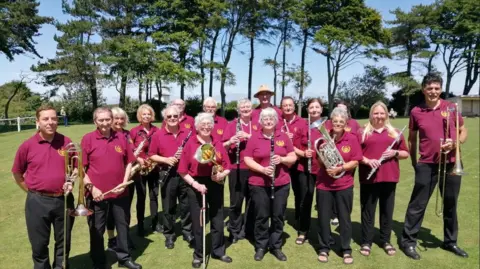 Band members wearing maroon polo shirts with black trousers holding brass instruments smiling while standing on grass near trees. 