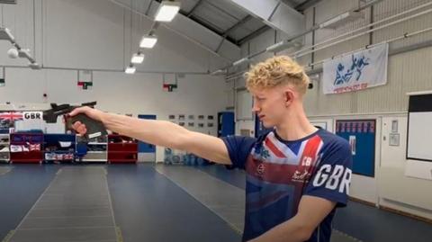 Man in blue union jack shirt holding a gun in a hall