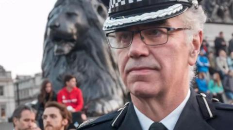 Julian Bennett wearing glasses and a police uniform in Trafalgar Square with crowds of people behind him