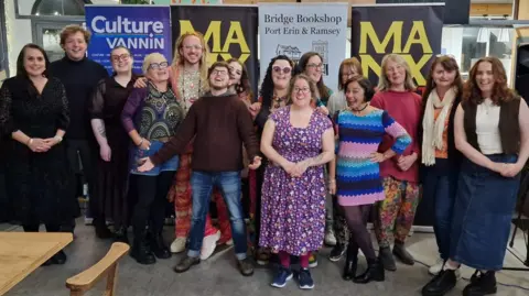 A group of men and women grouped together smiling wearing colourful casual clothes in front of banners that read Manx, Culture Vannin and Bridge Bookshop Port Erin & Ramsey. The people are all smiling and a man in the middle of the group is holding his arms stretched out and has his head tilted back.