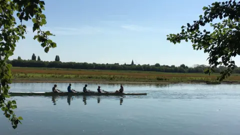 Wheatfields A rowing boat with four rowers and a cox move through the picture on the river in the sun with a meadow behind them and a glimpse of a church spire on the horizon