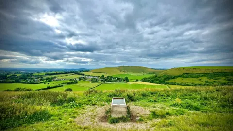 Moxley THURSDAY - The rolling green Dorset countryside is photographed from Compton Abbas. A metal water trough is in the centre of the frame, behind the countryside is dotted with trees and hedges with two hills. The sky is covered in grey skies
