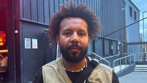 A man sits in the courtyard outside a music venue. He's got afro hair, a beard, and wears a light brown vest/gilet over a long-sleeved black top.
