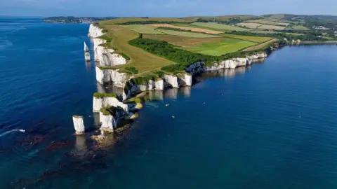 Gary Beach TUESDAY - An aerial photograph of the white cliffs at Old Harry Rocks on the Dorset coast. There are several stacks of cliff on this stretch of coast that stick out into the sea. Behind there are green frields and in the foreground the sea is a bright blue on a sunny day