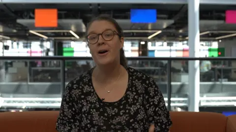 Catriona Aitken - who has glasses and tied-back brown hair and is wearing a black floral dress - sat on an orange sofa, with the colourful panels of the BBC Wales office behind her