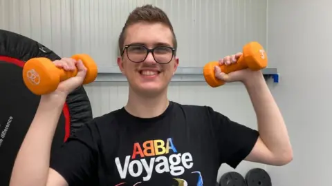 Zac Hollinshead, lifting two weights in a gym, and smiling 