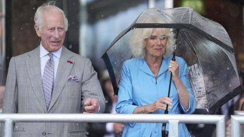 King Charles points and Queen Camilla in a light blue dress smiles while she holds an umbrella.