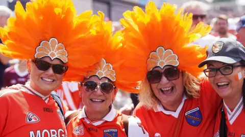 Three women wear bright orange feather hates. A forth is wearing a baseball cap