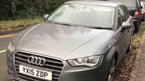 An abandoned grey 2015 Audi A3. It is parallel-parked at the side of a leafy road, with other cars parked up behind it
