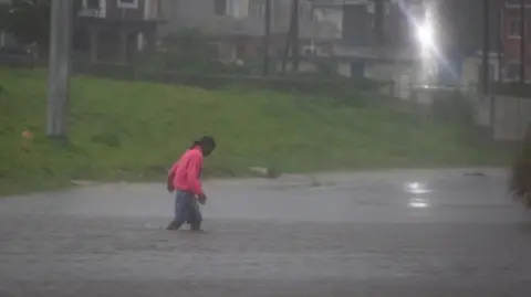 Reuters Man in bright pink jumper and shorts wades through flooded street