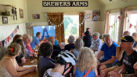 Dozens of people sit on chairs in a pub, watching a television that is showing the Olympic Games