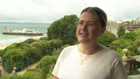 Councillor Millie Earl smiles at an unseen interviewer in an ornate garden above Bournemouth Pier. She wears glasses and a white short-sleeved top.