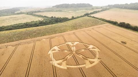 An aerial view of a crop circle resembling a five-spoked wheel with a rural landscape in the background