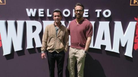Rob McElhenney and Ryan Reynolds in front of Welcome to Wrexham logo