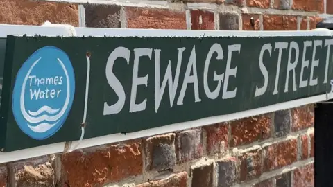 A hand-painted street sign reading 'Sewage Street' with the Thames Water logo next to it, with a brick wall behind it