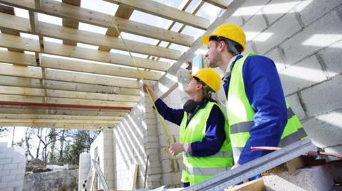 A man and woman wearing high-vis jackets and yellow hard hats hold a measuring tape in a house under construction