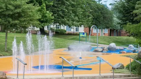 A splashpad with water spraying into the air, in front of trees and a grassy slope, with residential houses in the background