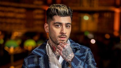Zayn, a man with his finger on his chin. His hand has many tattoos and he's wearing a navy blue blazer and white shirt