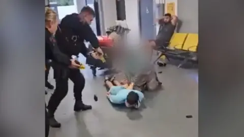 A police officer points a taser at a man lying face down on the floor, with a woman on her knees by his side. Another man is sitting on an airport bench behind them with his hands on his head.