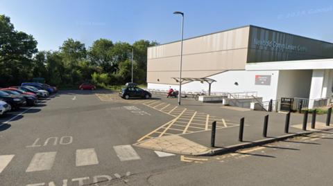 A brown building with a sign that says Newbold Comyn Leisure Centre, a full car park is also visible