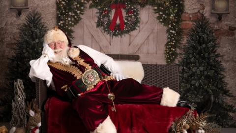 Allan Evans dressed as Santa, lying down on a sofar with his head propped on his hand and a Christmas tree in the background