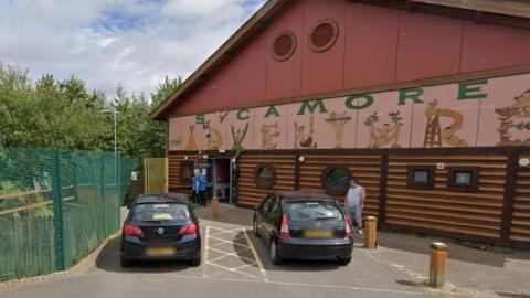 Two cars are parked outside a brown and red building. On the building are the words "sycamore adventure". The word "adventure is written by drawings of children spelling out the letters. A green fence surrounds the building