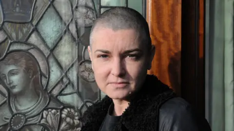 David Corio/Redferns/Getty Images Sinéad O'Connor - A woman with a shaved head looks at the camera with a closed-mouth smile in front of a stained glass window portraying religious imagery
