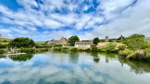 Philippa WEDNESDAY -  A body of blue water in the foreground with white clouds reflected. On the far bank are four traditional stone houses and a church tower. The blue sky is streaked with fluffy white clouds on a sunny day. The banks of the water are green with plants and trees.