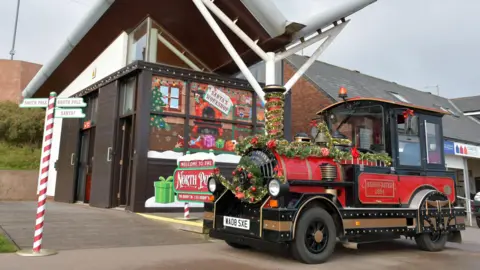 Bridlington's land train parked up at a station, it is red and black, with gold trim and decorated in tinsel and baubles. A red and white pole pointing left to the south pole and right to the north pole is in front of the train to create a festive feel