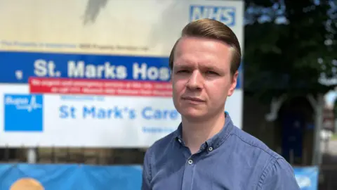 Joshua Reynolds in a blue shirt, standing in front of a NHS hospital sign