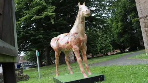 A brightly painted, life-sized sculpture of a horse stands on a patch of grass. Red handprints and birds are among the designs on the horse. 