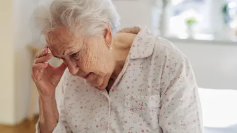An elderly woman in white pyjamas with a pink floral design sits on her bed, the background is out of focus, and she holds her hand to her head as if she's experiencing a headache or has forgotten something