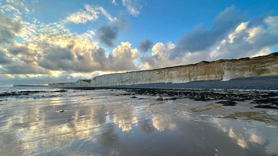 White chalk cliffs viewed from the sea, with a grey shingle beach. The clouds overhead are reflected in the water