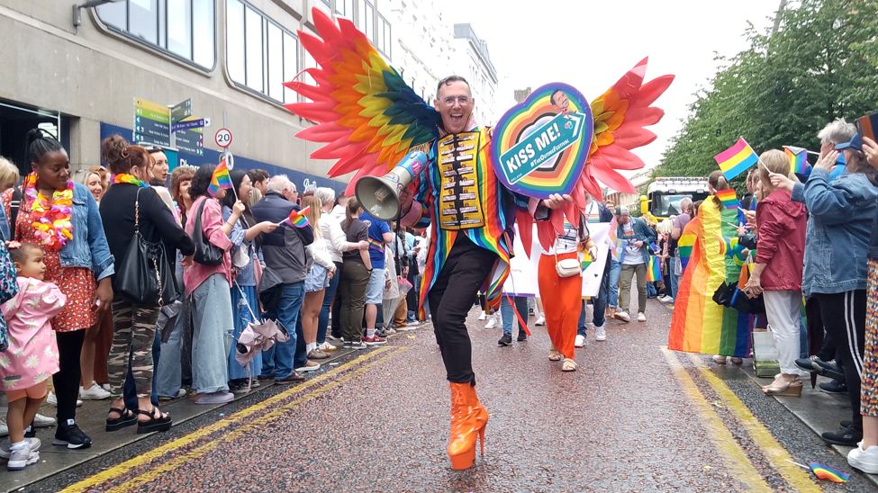 Person in large orange heels wearing a rainbow jacket and rainbow wings. They are carrying a megaphone and a sign that reads "kiss me"