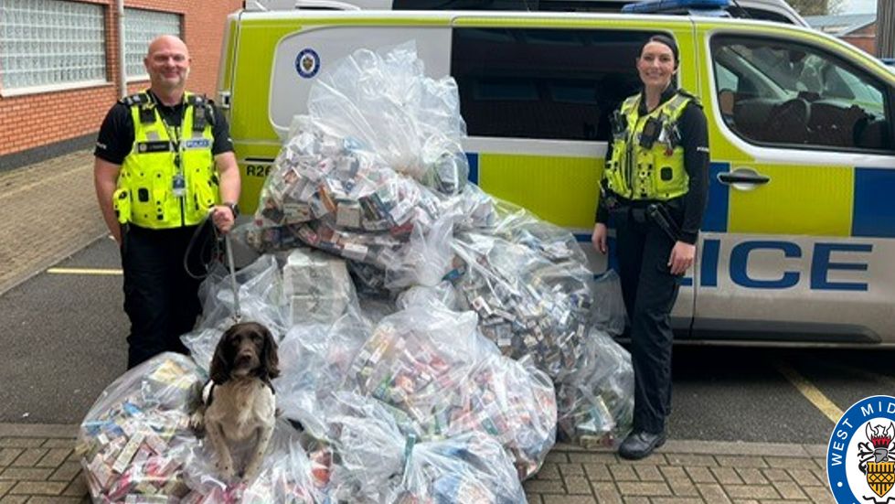 Police officers pictured wearing police uniform in front of a police van. The image includes plastic bags piled high and full of seized products, with a sniffer dog sat on the top of one of the bags. 