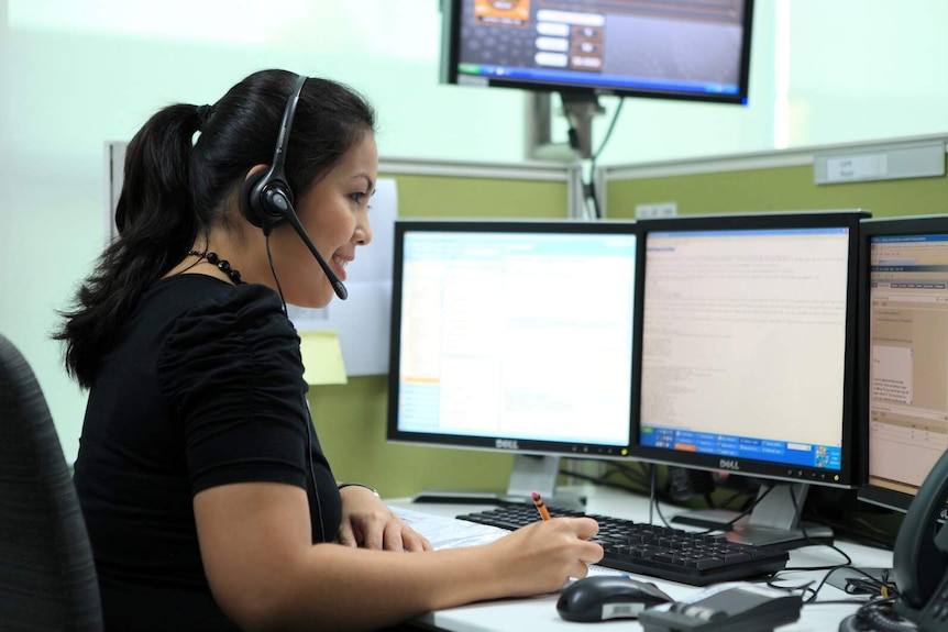 A woman smiles as she looks onto three computer screens while wearing a headphone set with a microphone.
