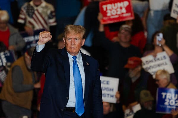 Donald Trump raises his right hand in a fist in front of a crowd, some of them holding Trump 2024 signs. He is wearing a dark suit, light blue shirt and blue tie.