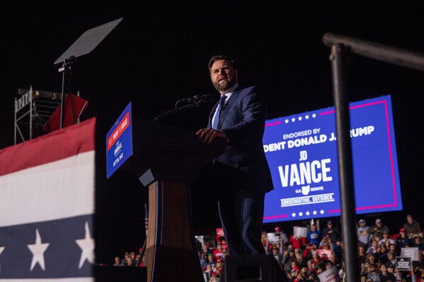 J.D. Vance standing on a stage addressing a crowd. He is wearing a beard and a dark blue suit. A sign behind him reads “Endorsed by President Donald J. Trump.”