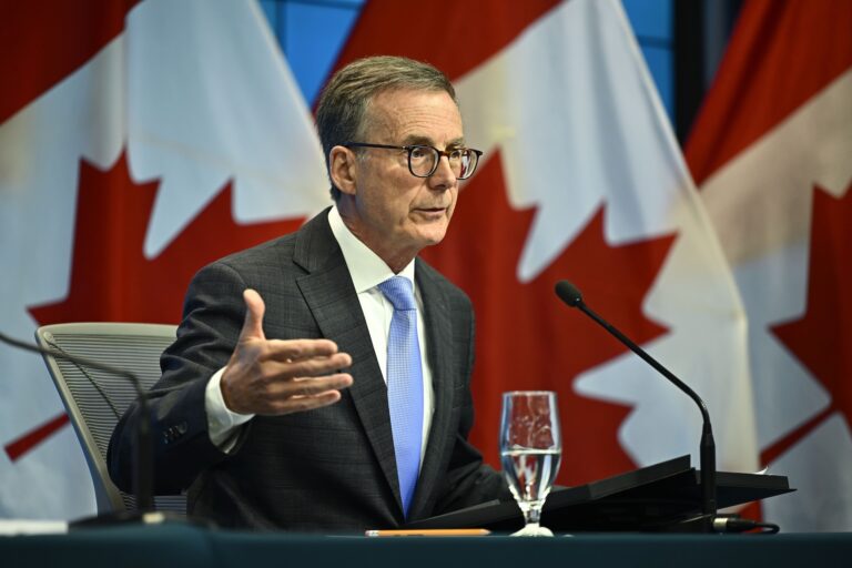 Bank of Canada governor Tiff Macklem speaking into a microphone and gesturing with his right hand while sitting at a table. Three red-and-white Canadian flags are seen behind him.