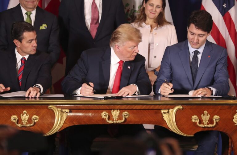 Donald Trump looks to his left as Prime Minister Justin Trudeau signs the document; Mexico's president at the time, Enrique Pena Nieto, looks over from Trump's right. The three leaders are seated at a wooden table with gold ornamentation.