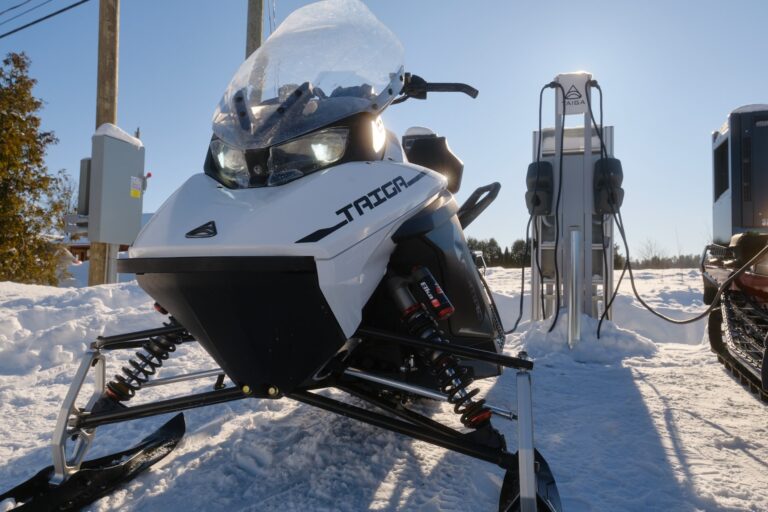 A white snowmobile labeled "Taiga" is parked in front of an electric charging station on a snowy landscape with a clear blue sky.