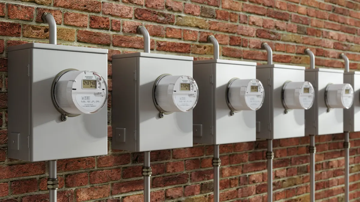 A row of five, silver-colored electric meters on a red brick wall.